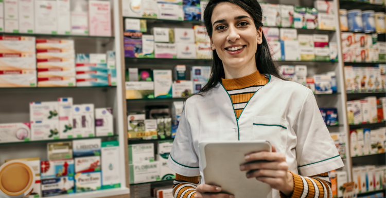 female pharmacist holding a tablet computer, standing in front of shelves of medication.