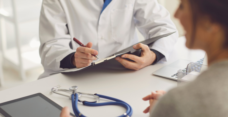 A person sitting at a desk holding a clipboard and pen. A stethoscope is on the desk. Sitting opposite them is a female.