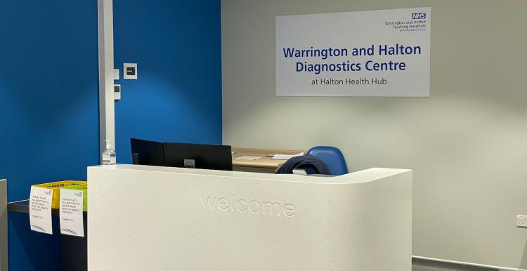 The reception area for Halton CDC. Image shows a large white reception desk with a sign on the wall behind it reading, 'Warrington and Halton Diagnostic Centre at Halton Health Hub'