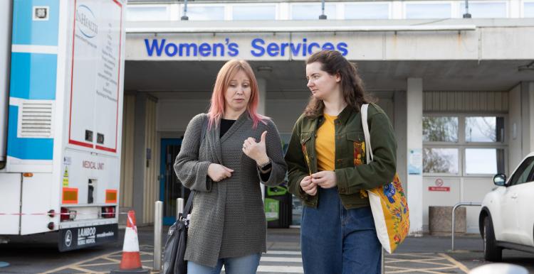 Two women chatting as they walk away from a building in the background. On the building is a sign which reads, 'Women's Services'