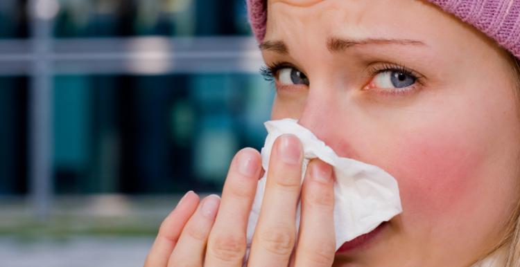 face of a woman wearing a pink wooly hat, she is looking into the camera and wiping her nose with a paper tissue