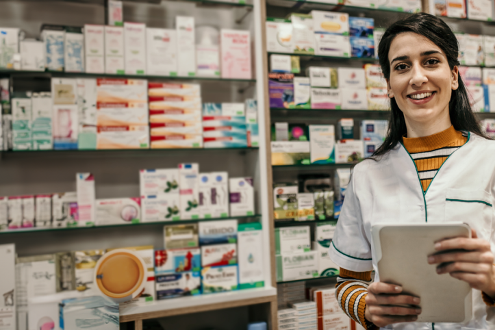 female pharmacist holding an iPad standing in front of shelves of medication