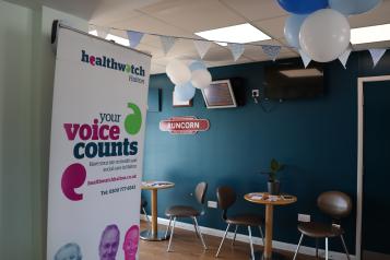 Healthwatch banner and two tables and chairs in the community lounge at Runcorn Train Station 