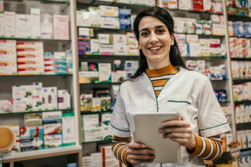 female pharmacist holding a tablet computer, standing in front of shelves of medication.