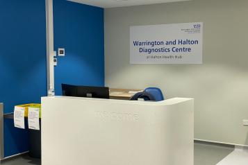 The reception area for Halton CDC. Image shows a large white reception desk with a sign on the wall behind it reading, 'Warrington and Halton Diagnostic Centre at Halton Health Hub'