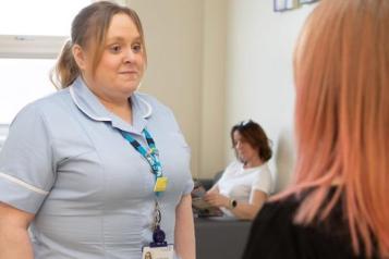 This image shows a healthcare worker in a light blue uniform, standing and speaking with a woman whose back is to the camera. The healthcare worker is wearing a lanyard with identification and looks engaged in conversation. In the background, another person is sitting on a couch, looking at something, possibly a phone or tablet. The setting appears to be a hospital waiting room