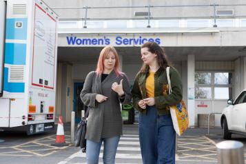 Two women chatting as they walk away from a building in the background. On the building is a sign which reads, 'Women's Services'