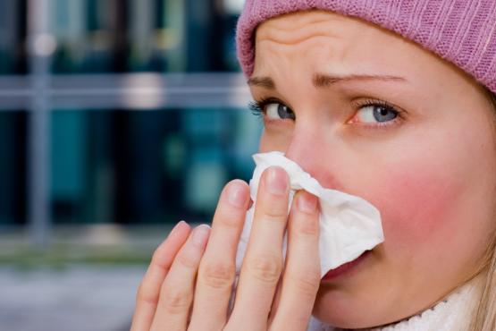 face of a woman wearing a pink wooly hat, she is looking into the camera and wiping her nose with a paper tissue